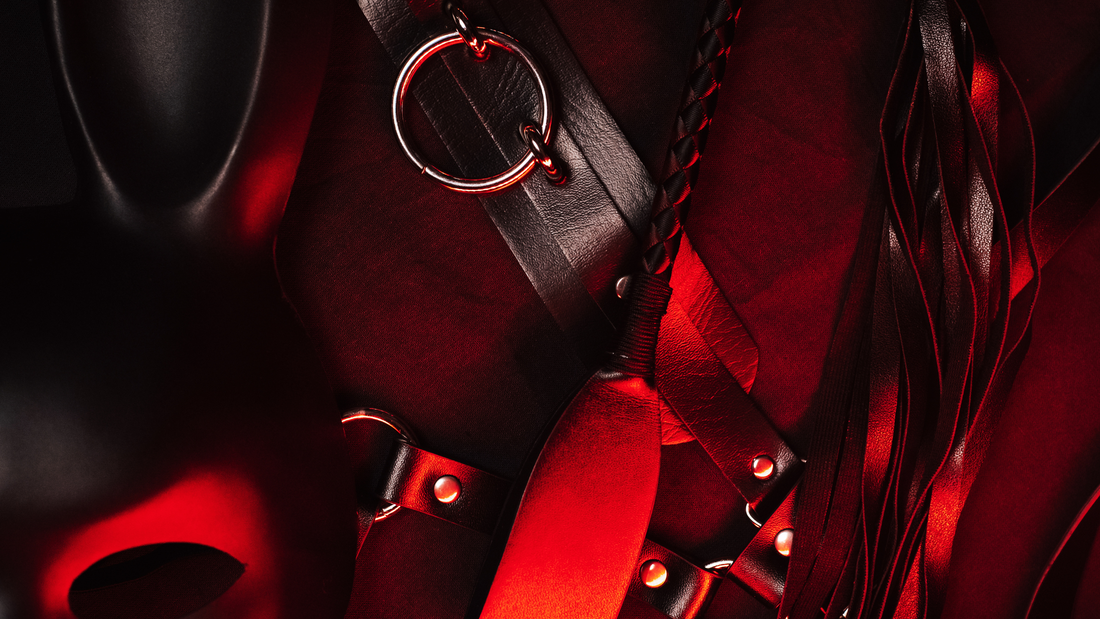 Beginner BDSM kit: mask, soft cuffs, paddle and flogger on a dark red background.