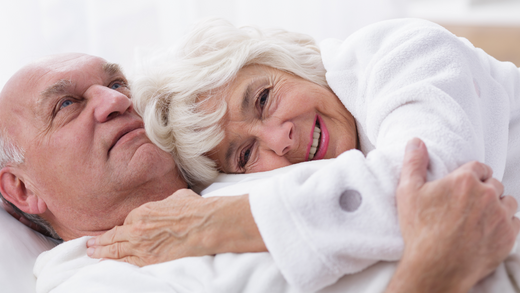 An elderly couple smiling and embracing each other while lying in bed, both wearing white bathrobes. The man is looking at the woman affectionately, and the woman is gazing at the camera with a joyful expression.