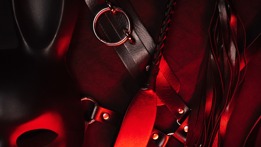 Beginner BDSM kit: mask, soft cuffs, paddle and flogger on a dark red background.