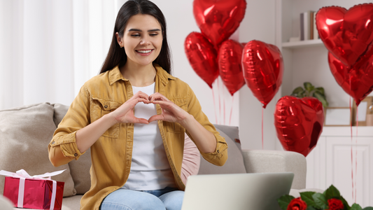 Smiling woman sitting on a sofa making a heart shape with her hands during a video call, surrounded by red heart-shaped balloons and a gift box, symbolising love and connection on Valentine's Day.