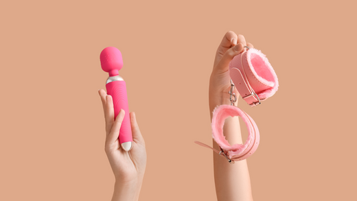 Two hands against a plain peach background, each holding different objects related to BDSM. The left hand holds a pink vibrator with a flexible neck and a bulbous head, designed for stimulation.